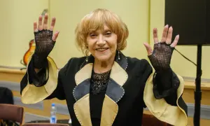 Uśmiechnięta starsza kobieta w czarnej marynarce z ozdobnym kołnierzem, w siatkowych rękawiczkach, unosi ręce.