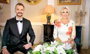 Teresa Werner i jej mąż, elegancko ubrani, siedzą przy bukiecie kwiatów.