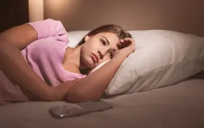 Młoda kobieta leży w łóżku, zamyślona. Telefon obok. Czy to dlatego popularna influencerka sypia tylko przy całkowitym zaciemnieniu?