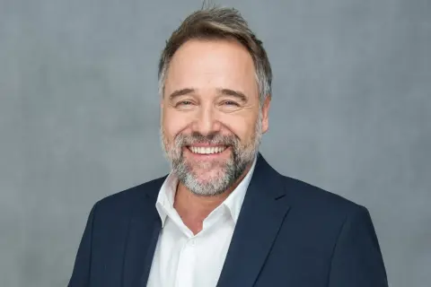 Uśmiechnięty Łukasz Nowicki w garniturze. Ile lat ma Łukasz Nowicki? Wiek nie jest widoczny na zdjęciu, ale wygląda na dojrzałego mężczyznę.