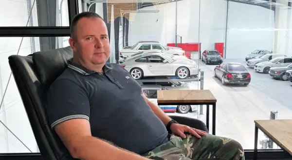 Robert Michalski, w wieku około 40-50 lat, siedzi w czarnym fotelu. W tle widać kolekcję samochodów.