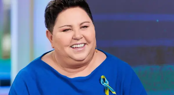 Uśmiechnięta Dorota Wellman w niebieskim swetrze z przypinką w barwach Ukrainy. Jej wykształcenie i doświadczenie pomagają jej w pracy.