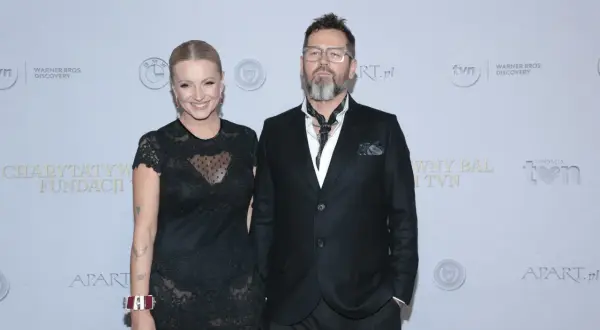 Michał Cessanis żona, ubrana w koronkową sukienkę, pozuje obok męża w eleganckim garniturze.