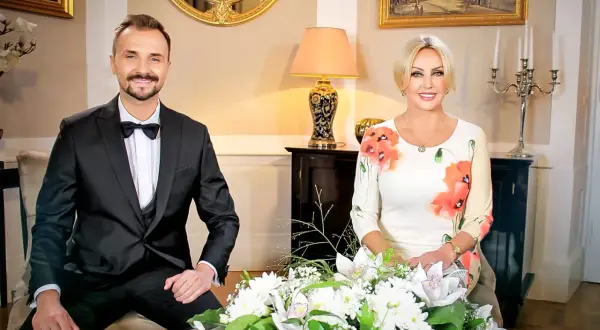 Teresa Werner i jej mąż, elegancko ubrani, siedzą przy bukiecie kwiatów.