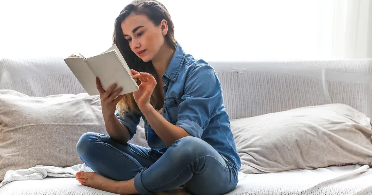 Odkryj sekrety domowych bibliotek gwiazd! Co naprawdę czytają znani i lubiani? Młoda kobieta w dżinsowej koszuli zanurzona w lekturze na kanapie.