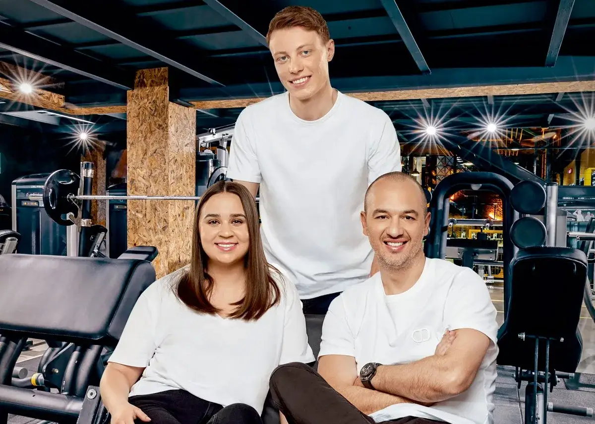 Trzy uśmiechnięte osoby w białych koszulkach na tle siłowni. Czy partner znanej trenerki fitness rzeczywiście sam odniósł sukces?