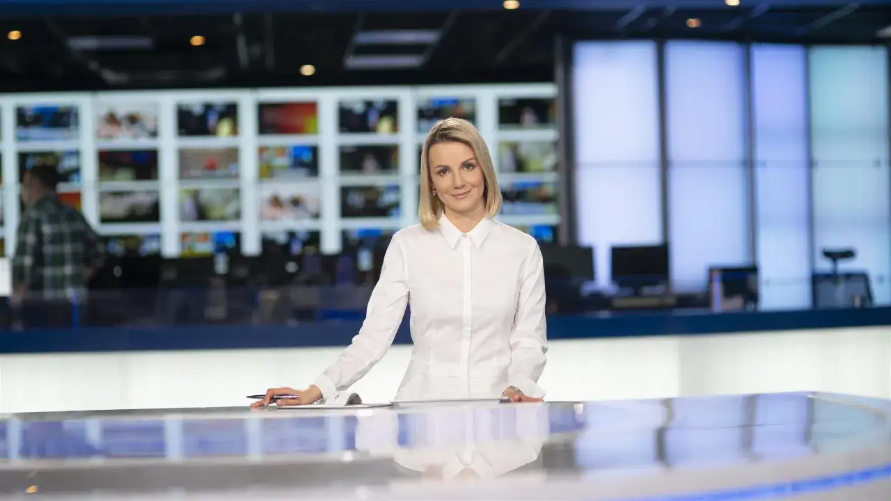 Znana dziennikarka w białej koszuli, za nią ekrany z wiadomościami. Czy to odpowiedź na pytanie, dlaczego znana dziennikarka przeszła do konkurencji bez pożegnania z widzami?
