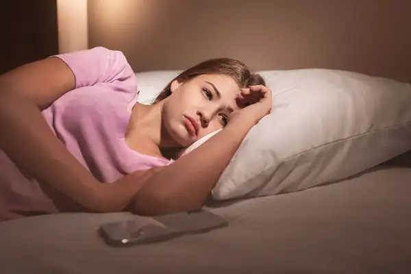 Młoda kobieta leży w łóżku, zamyślona. Telefon obok. Czy to dlatego popularna influencerka sypia tylko przy całkowitym zaciemnieniu?