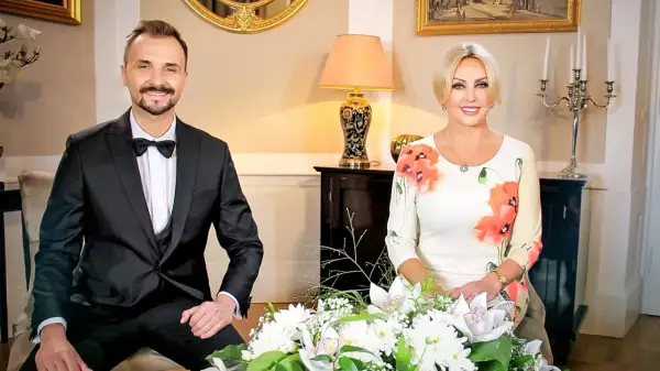 Teresa Werner i jej mąż, elegancko ubrani, siedzą przy bukiecie kwiatów.