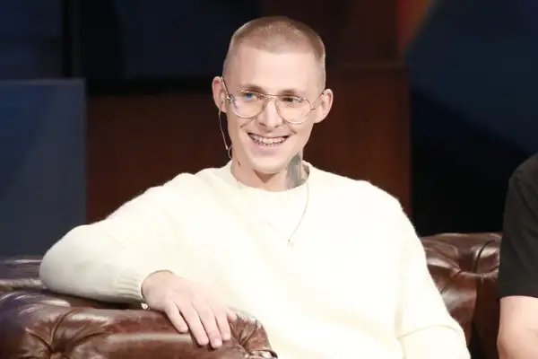Młody mężczyzna z tatuażami na szyi, w okularach i swetrze, uśmiecha się. Mateusz Trąbka wiek nieznany, ale wygląda na młodego.